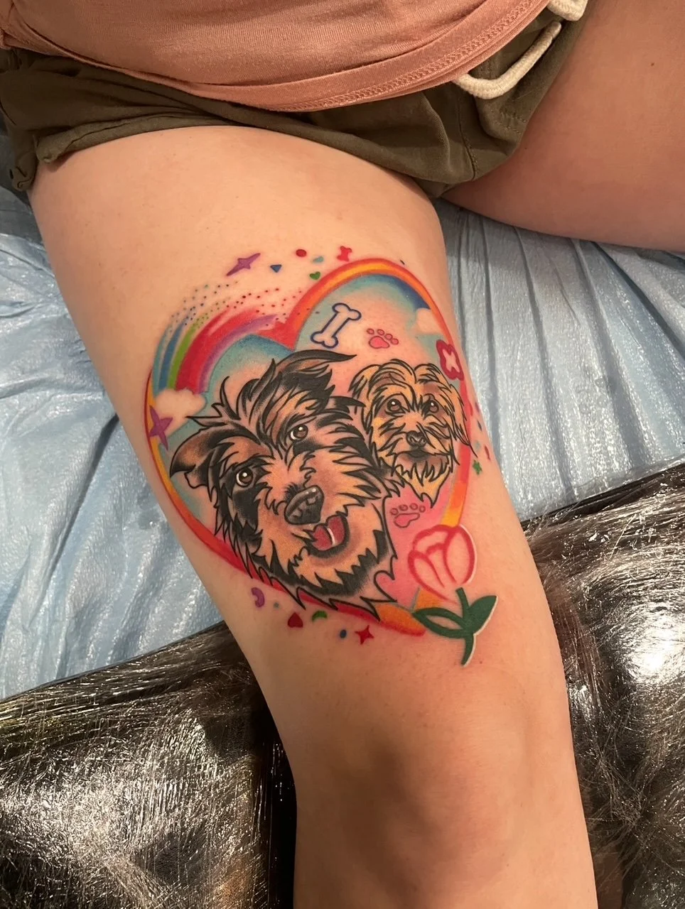 Colorful tattoo on person's thigh of a heart-shaped rainbow with two realistic dog faces inside, surrounded by small stars, hearts, paw prints, a bone, and a pink flower.