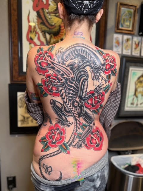 A woman with their back facing the camera, showcasing a xenomorph tattoo surrounded by red roses and green leaves.