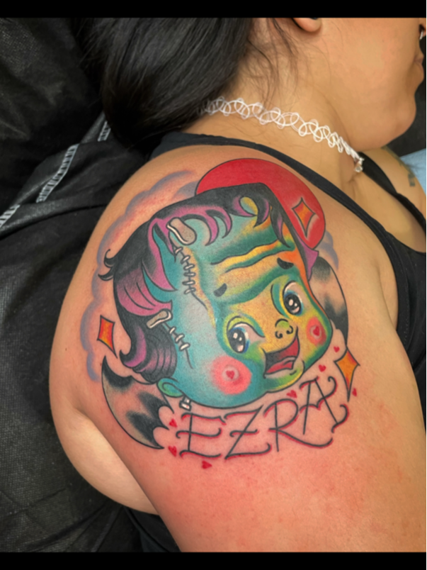 Colorful new school tattoo of a cartoonish girl's face with rainbow-colored hair, earrings, and blushing cheeks on a person's upper arm, with the name 'EZRA' and small red hearts and diamonds surrounding it.