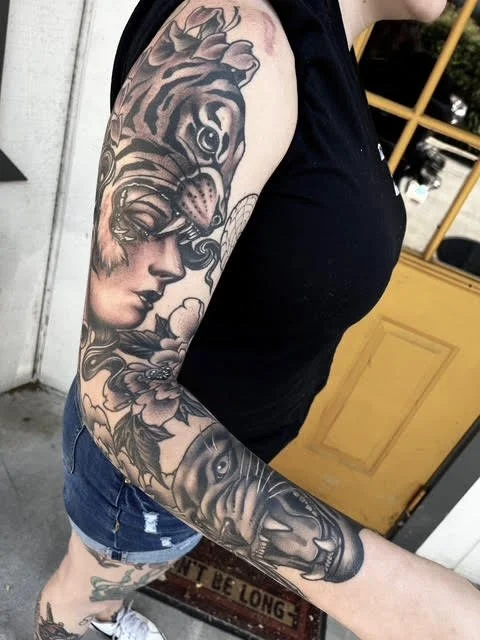 Tattooed arm featuring a woman’s face, a tiger’s face, and a large eye, with flowers and clouds surrounding the designs.