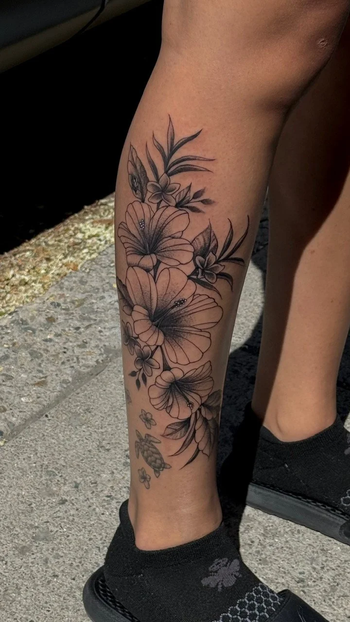 A person's lower leg with a floral tattoo featuring large flowers, leaves, a butterfly, and a small turtle, in black and gray ink.