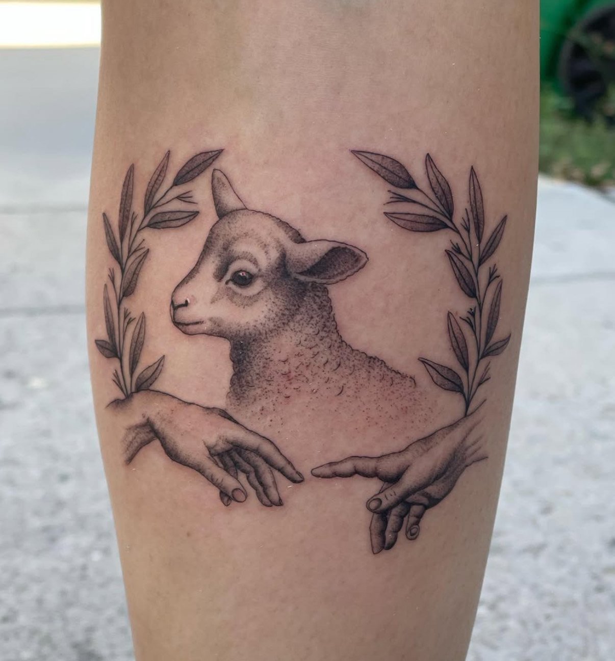Tattoo of a lamb's head surrounded by a laurel wreath, with two hands reaching towards each other below.