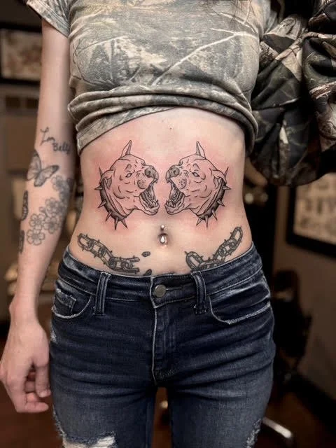 Tattoo of two fierce dog heads facing each other on the person's lower abdomen, with a chain and bullet tattoo below the navel.