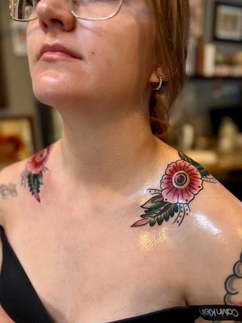 A person with glasses and tattoos on their shoulder and neck, wearing a black top, in a tattoo shop in Carson City.
