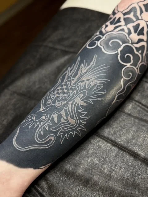 Tattoo of a dragon on a person's calf, featuring detailed lines and cloud patterns in white ink over a blackout sleeve