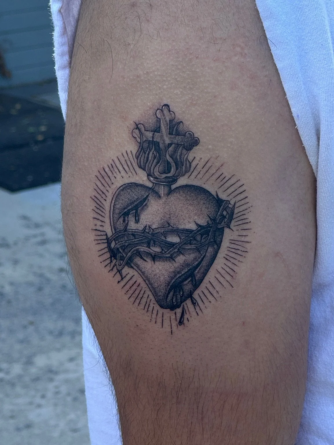 A black and gray tattoo of a broken heart with a crown of thorns and a small cross on top, surrounded by radiant lines.
