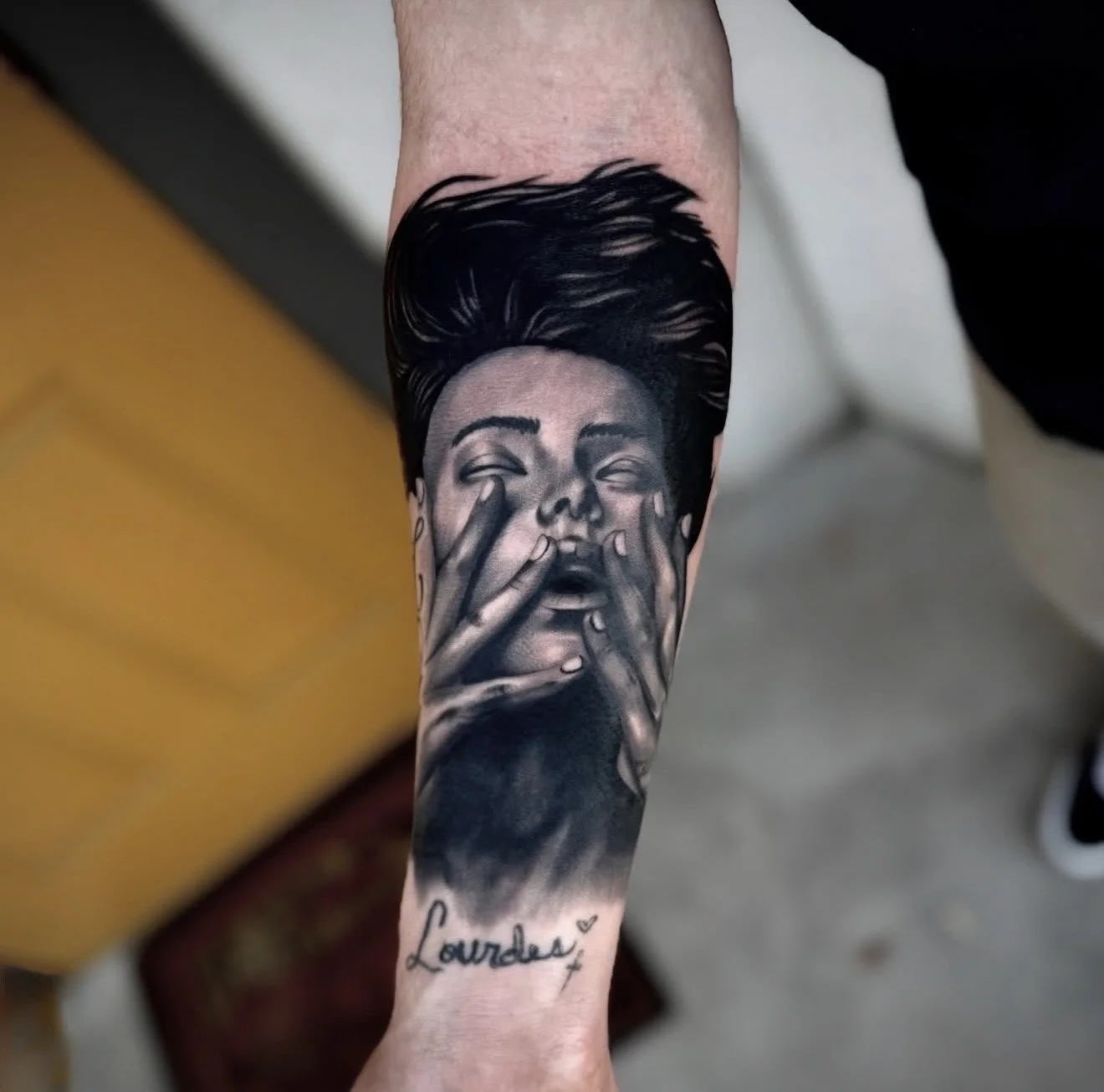 Black and gray tattoo of a woman with closed eyes and hands covering her face, with the name 'Lourdes' written below.