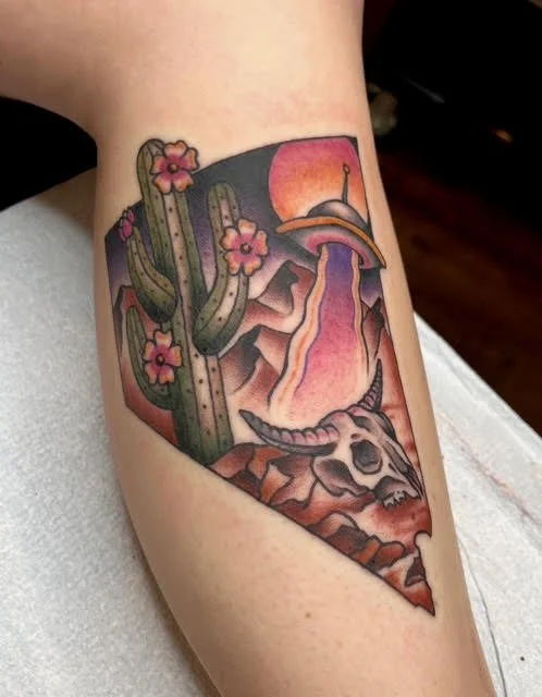 Tattoo of a nevada desert landscape with cactus, pink flowers, mountains, a waterfall, and a sunset, all enclosed in a frame.