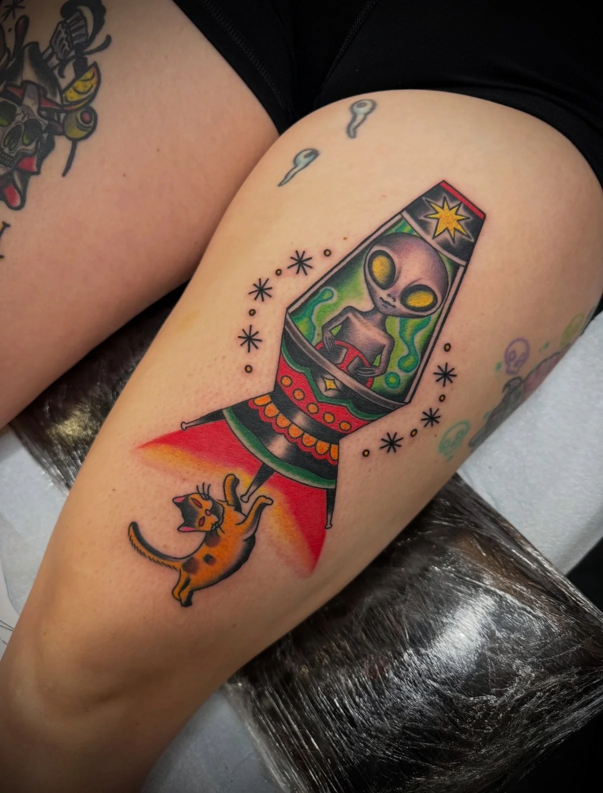 Tattoo of a cartoon-style alien in a spaceship with a cat below, surrounded by small star-like designs.