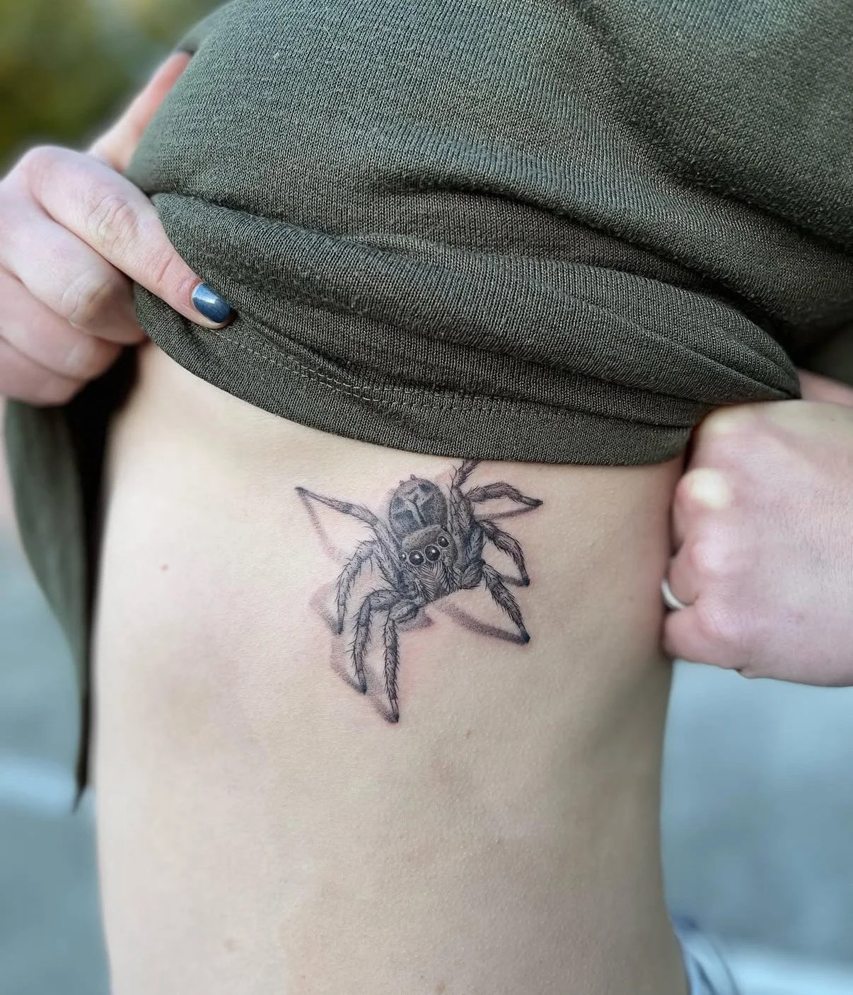 Tattoo of a spider on a person's side, partially concealed by the green shirt they are lifting.
