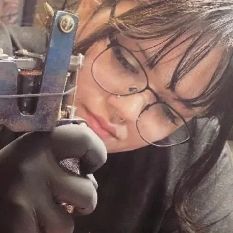 A female tattoo artist with glasses and nose piercings working closely with machinery, wearing gloves.