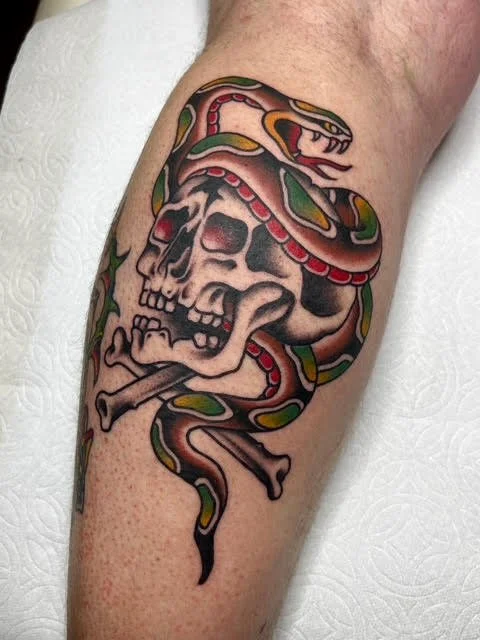 Tattoo of a skull with a snake wrapping around it and holding a dagger in its mouth.