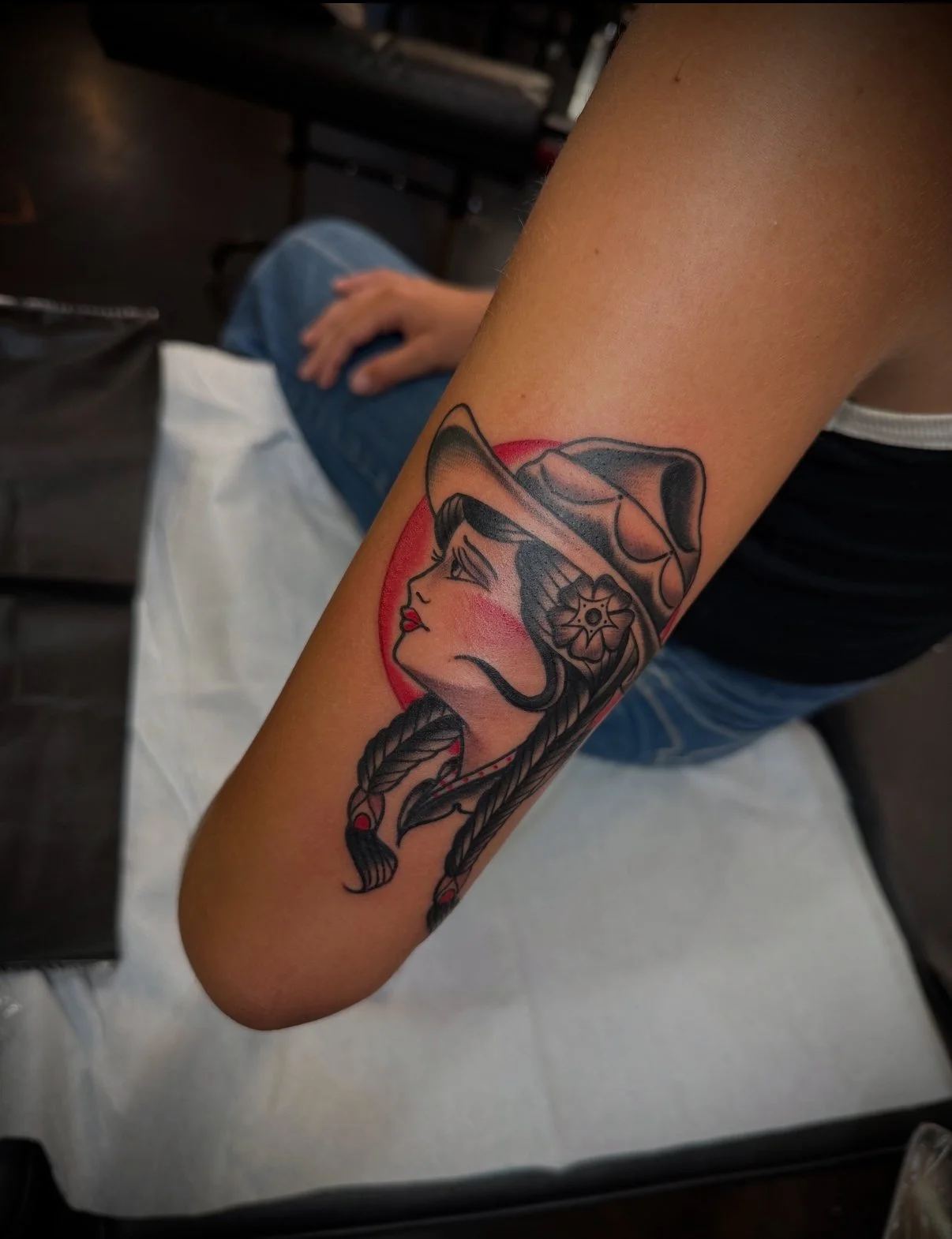 Tattoo of a woman with braided hair, a hat, and a flower, with a red circle background.