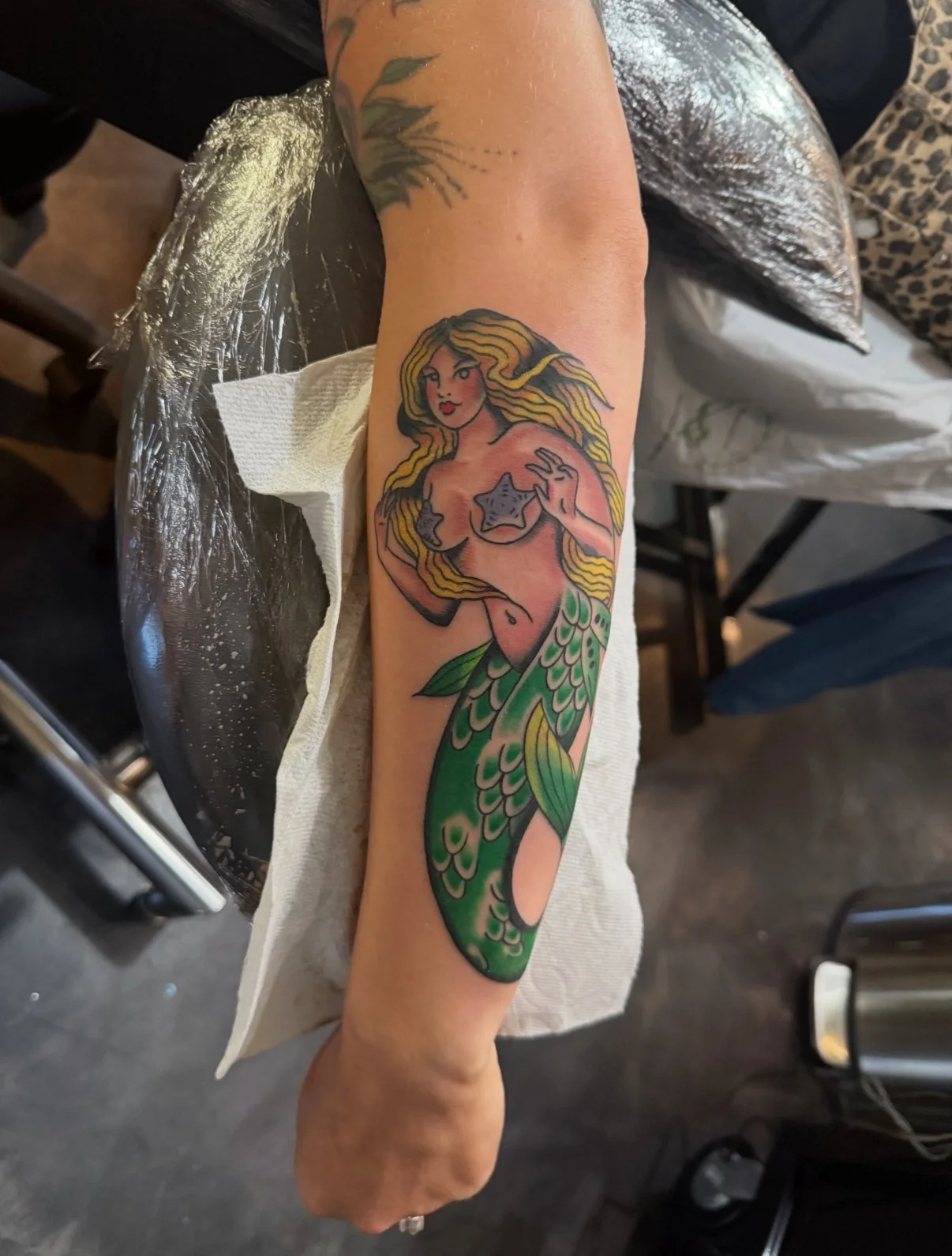 A colorful tattoo of a mermaid with long flowing blonde hair, holding her chest with one hand, on her arm resting on a table in a tattoo studio.