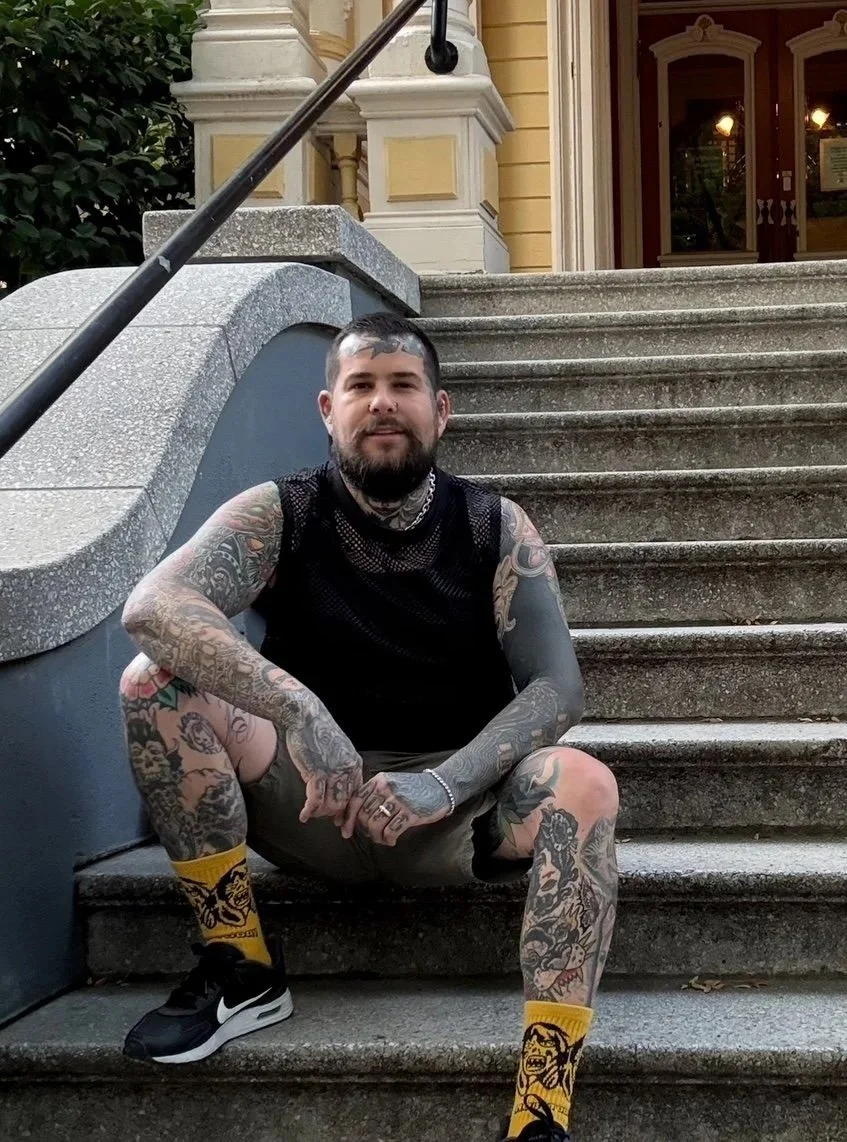 A tattoo artist sitting on outdoor stairs in front of a building with a yellow facade and red door, wearing black Nike sneakers and yellow socks with black skull designs.
