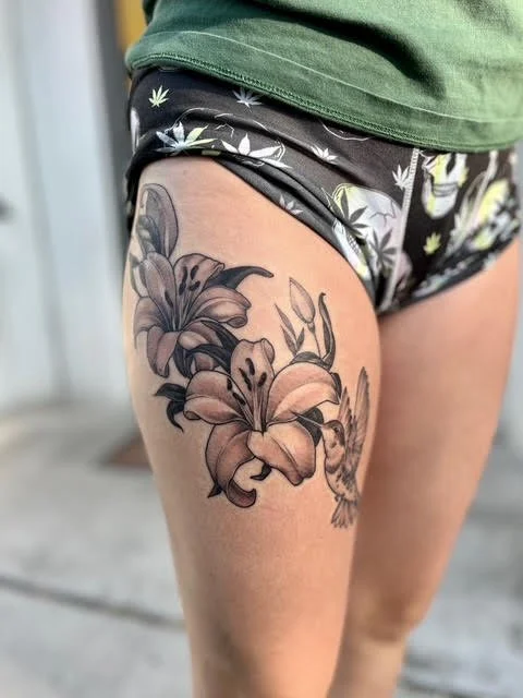 Tattoo of large lilies and a hummingbird on a person's thigh, wearing camouflage shorts and a green shirt.