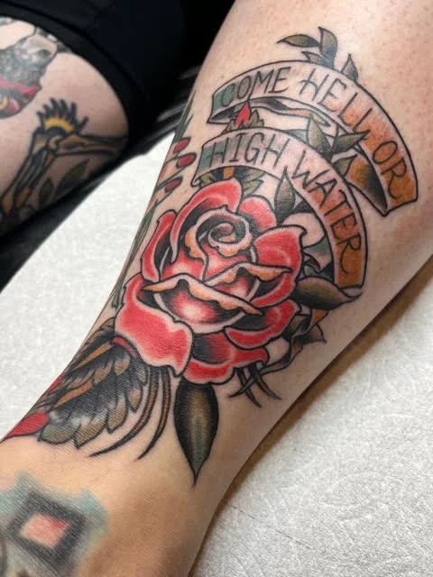 A tattoo on a person's arm featuring a red rose with green leaves, a banner with the words "Come hell or high water," and other decorative elements.