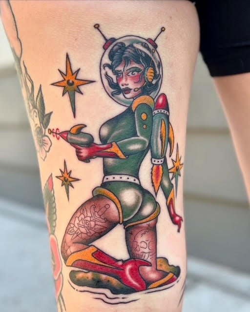 Tattoo of a female astronaut with a vintage space helmet and a power suit, holding a laser gun, surrounded by stars and planets, on a person's thigh.