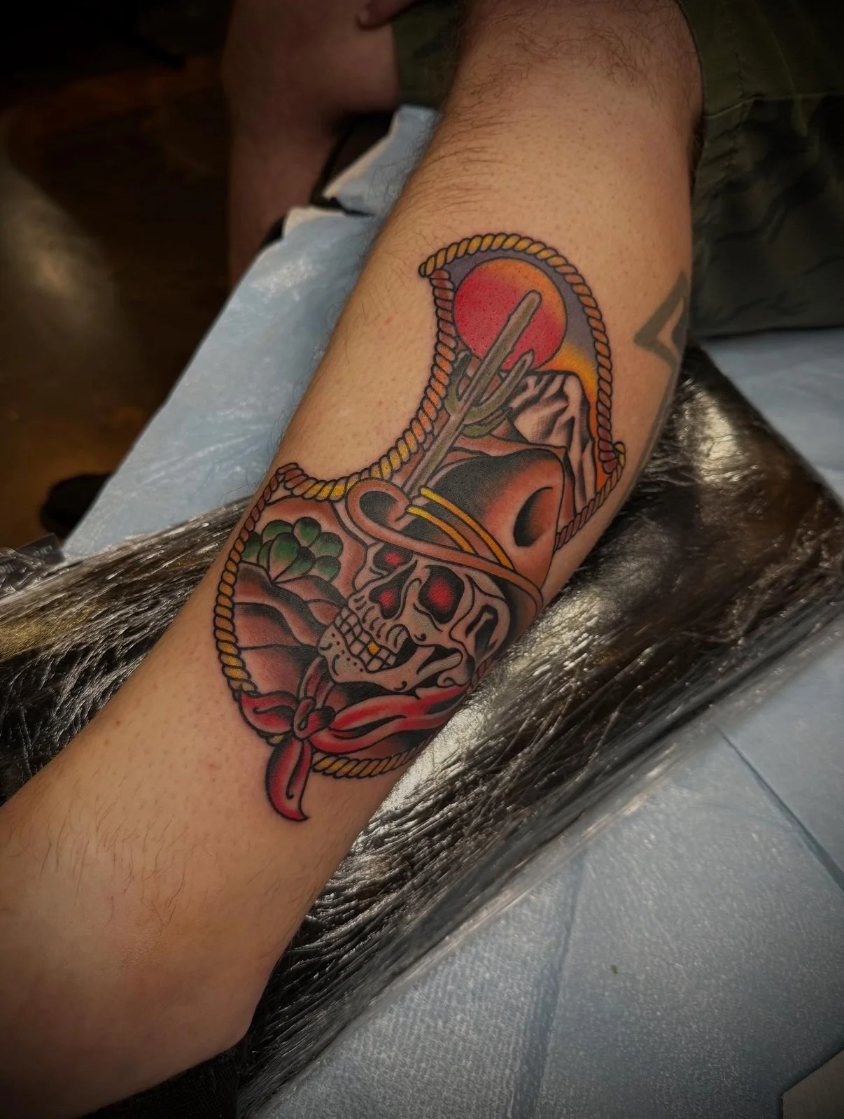 Colorful tattoo on person's forearm depicting a skull, cactus, mountain, sun, rope, and a red bandana.