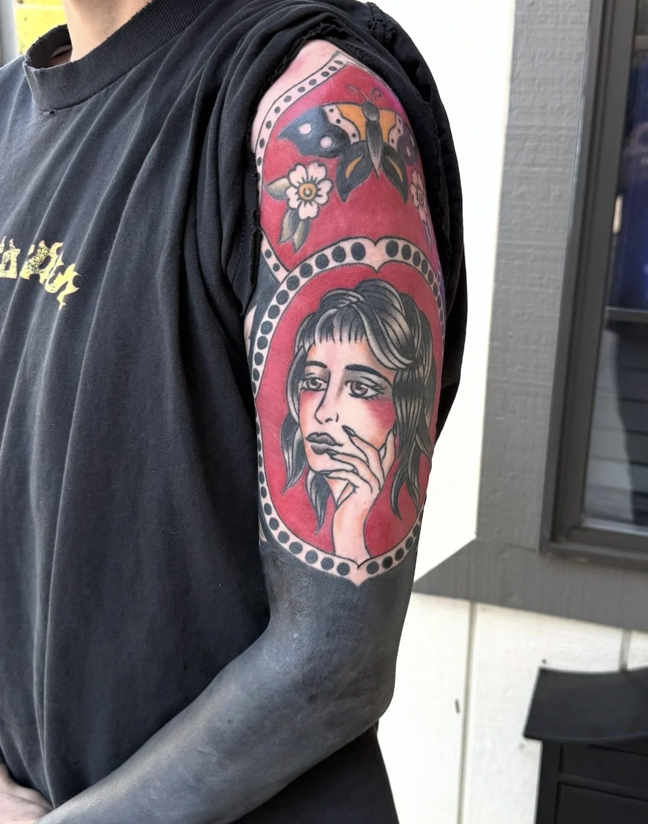 Tattoo of a woman's face with hand near her chin, surrounded by a red background and dotted border, on person's upper arm. Above it, a tattoo of a butterfly and flowers on the shoulder.