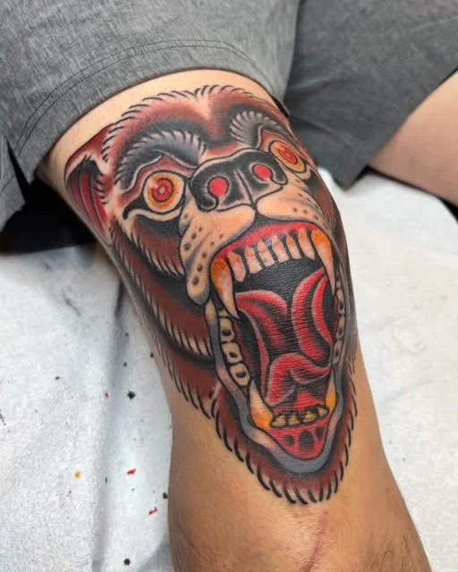 Tattoo of a roaring bear's face on a person's knee with detailed fur, eyes, and open mouth revealing teeth and tongue.