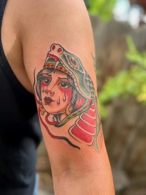 Tattoo of a woman with tears on her face, wearing a headdress that resembles a snake or dragon, on a person's upper arm.