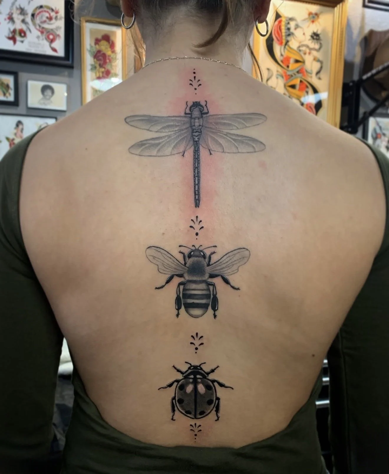Tattoo of three insects on a person's back, including a dragonfly at the top, a bee in the middle, and a ladybug at the bottom, with decorative dots and lines between them.
