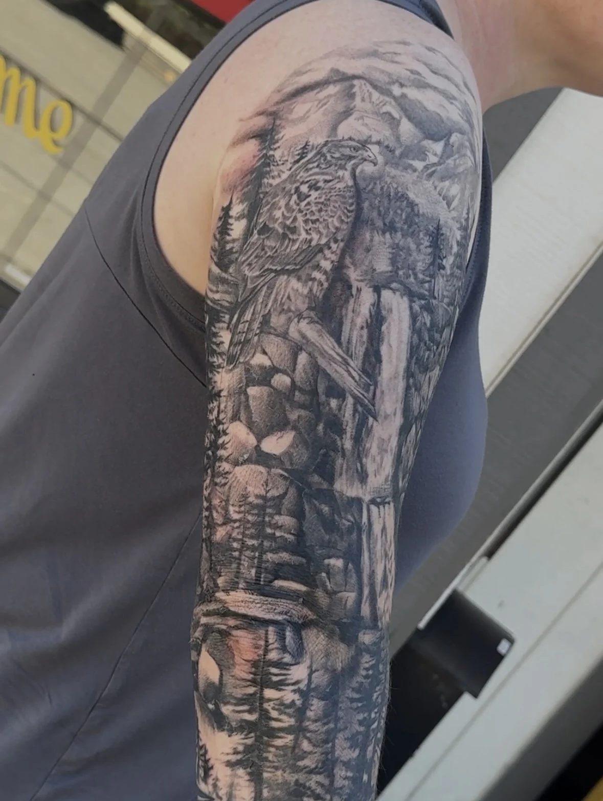 Black and gray mountain landscape tattoo featuring a mountain, trees, and a bird on the upper arm