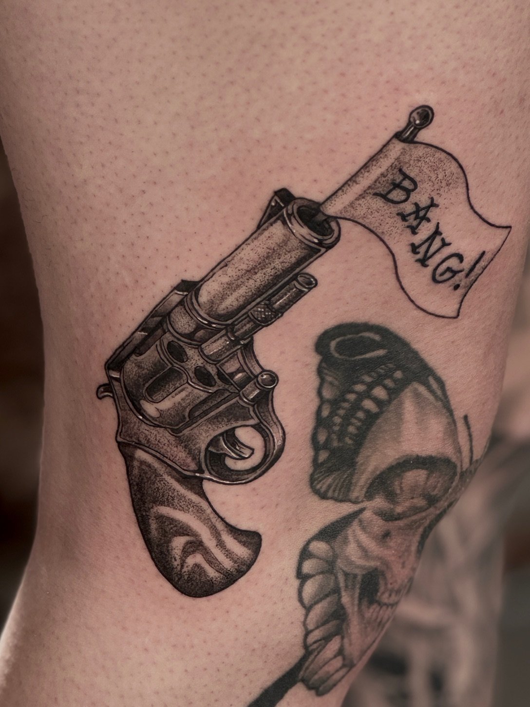 Tattoo of a revolver with a flag reading 'bang' and a skull with a snake wrapped around it.