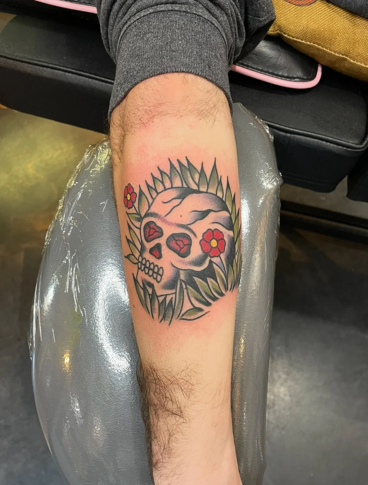 A tattoo of a skull with heart-shaped eye sockets, surrounded by green leaves and red flowers, on a person's forearm.