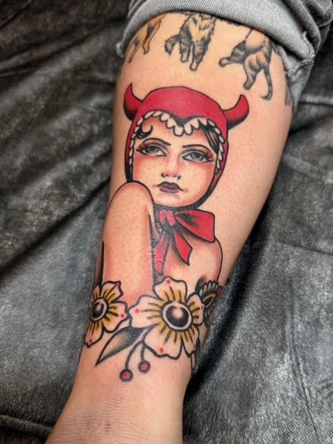 Tattoo of a woman with a retro style, wearing a red devil hat and a red ribbon, surrounded by sunflowers.