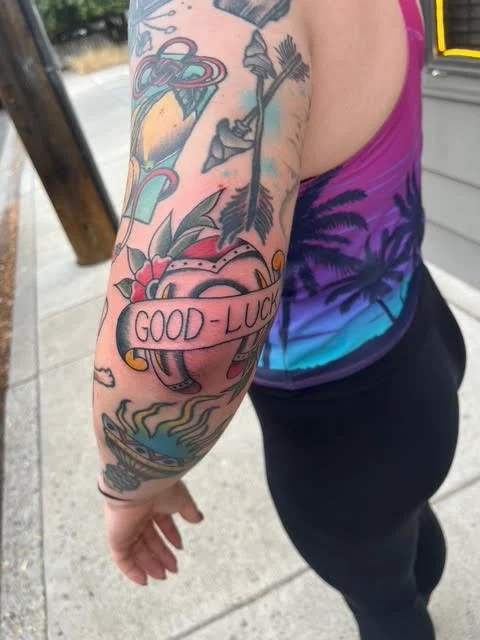 Close-up of a person's tattooed arm showing colorful traditional-style tattoos, including a heart with the words 'GOOD LUCK' on a banner.