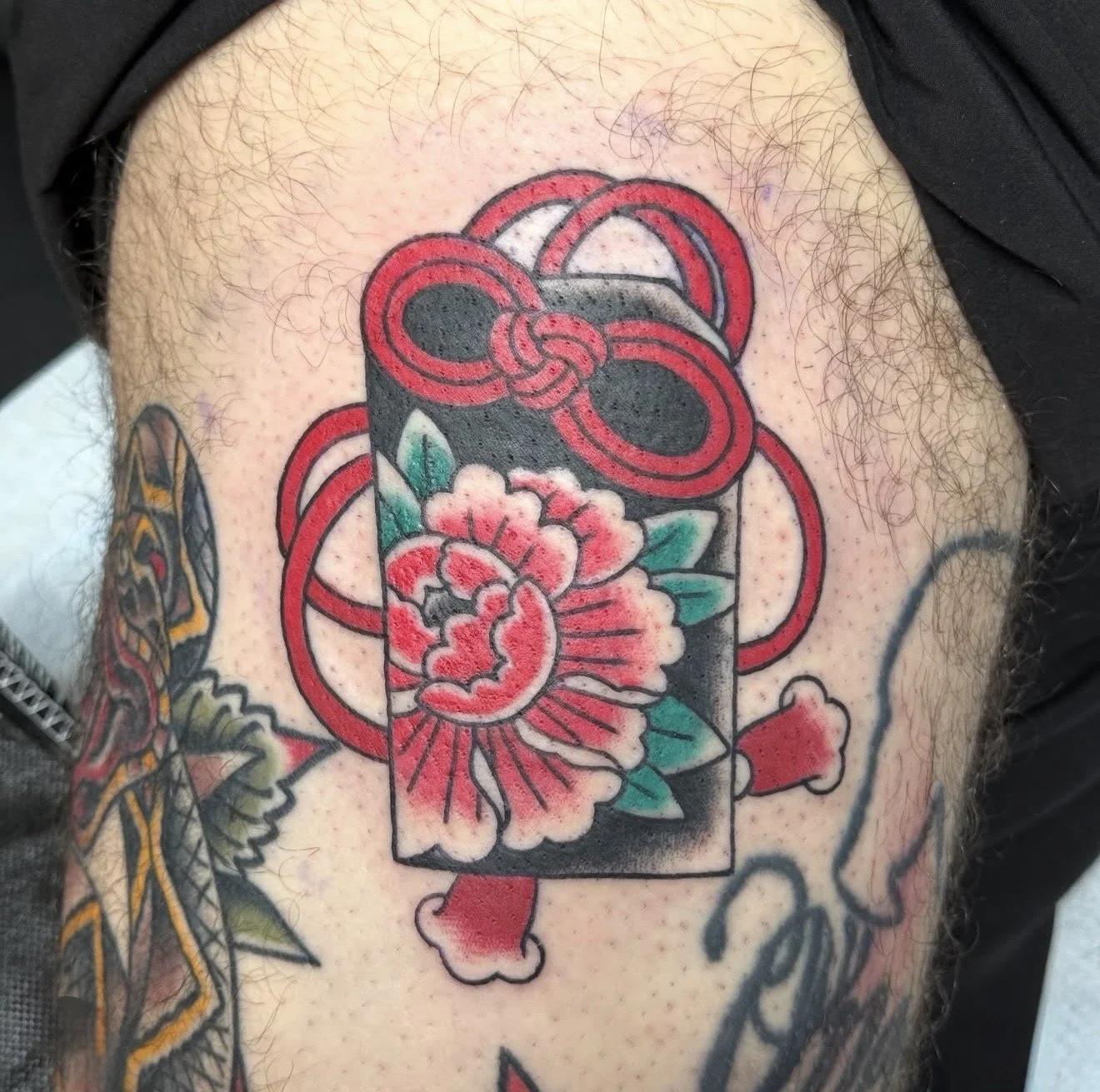 Traditional-style tattoo of a black can with a red and pink peony flower, green leaves, and red ribbon accents on a person's arm.