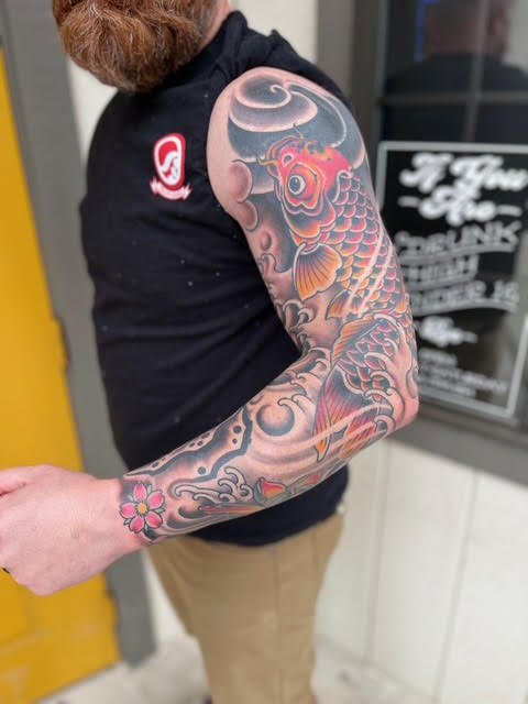 A man with a red beard and short hair showing off a colorful Japanese-style koi fish tattoo on his arm.