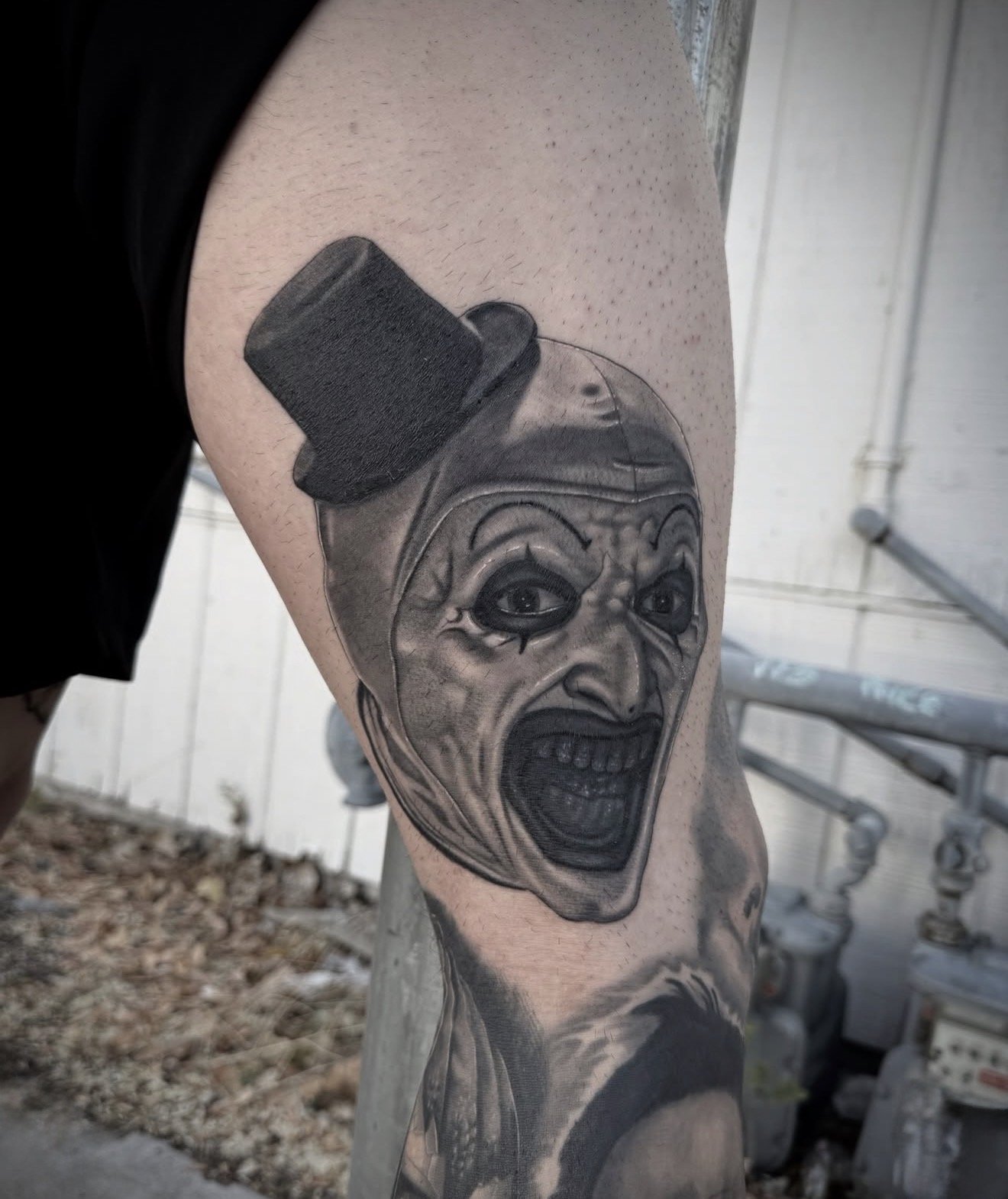 Black and gray realism tattoo of the terrifier exaggerated facial features, wearing a small black top hat, on a person's upper arm.