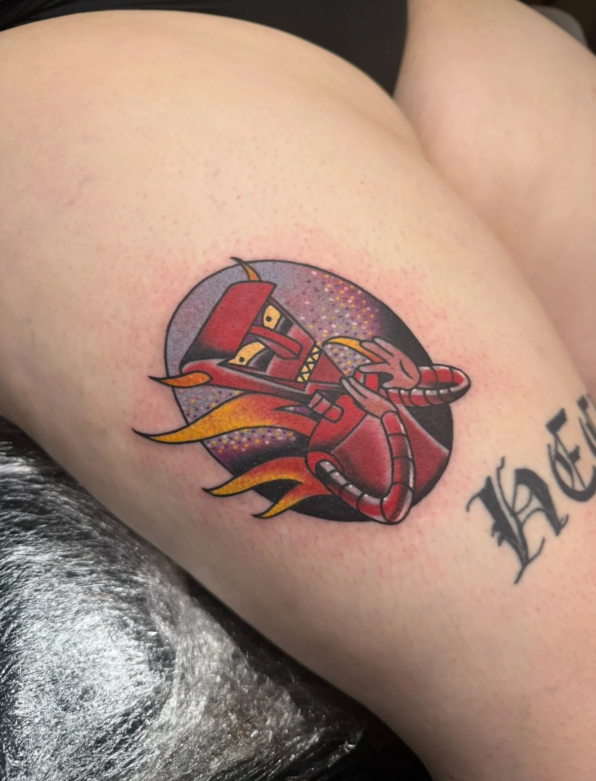 Tattoo of a cartoon-style red robot with flames in the background on a person's upper arm.