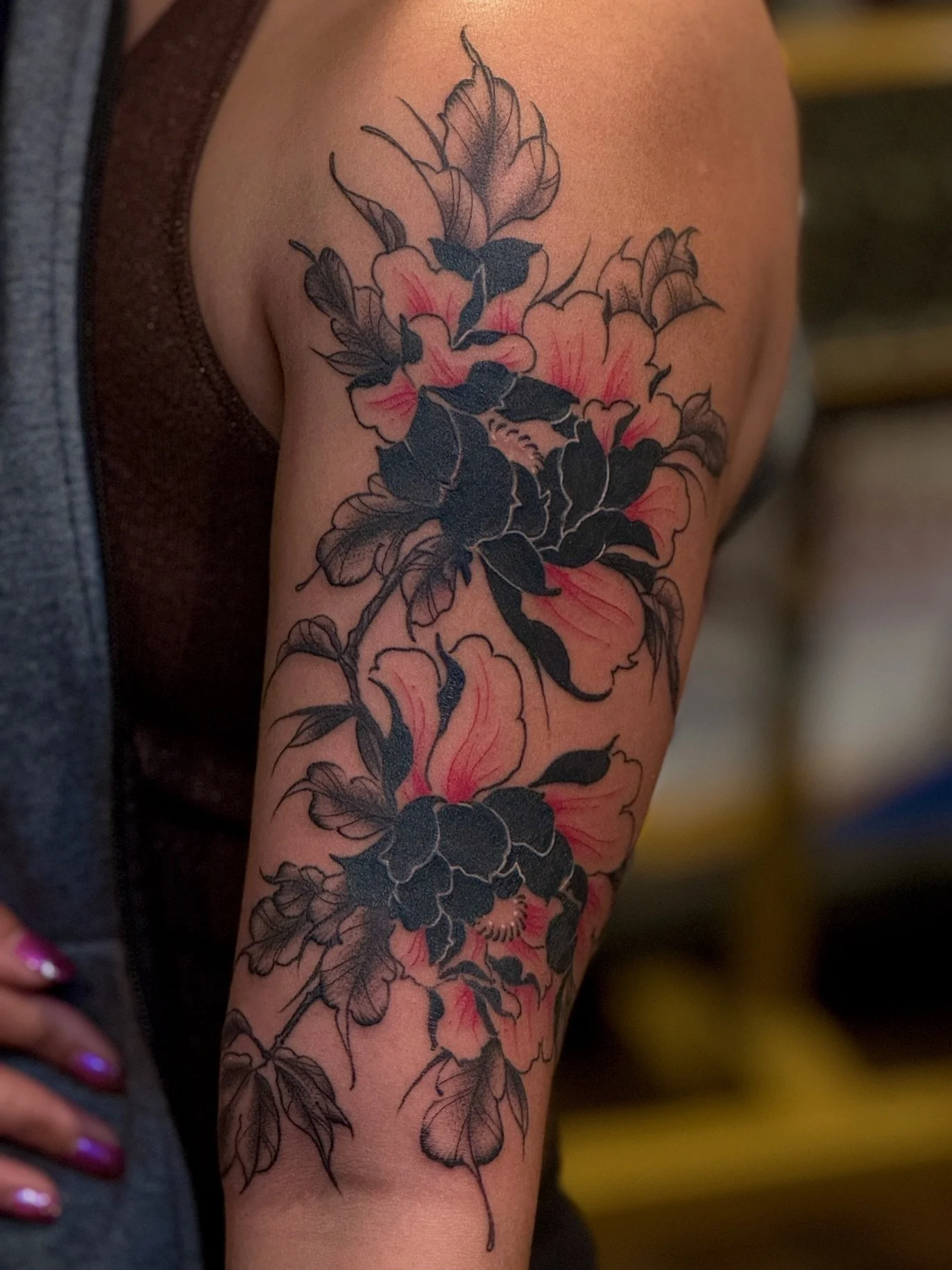 Tattoo of large pink and black flowers with leaves on person's upper arm.