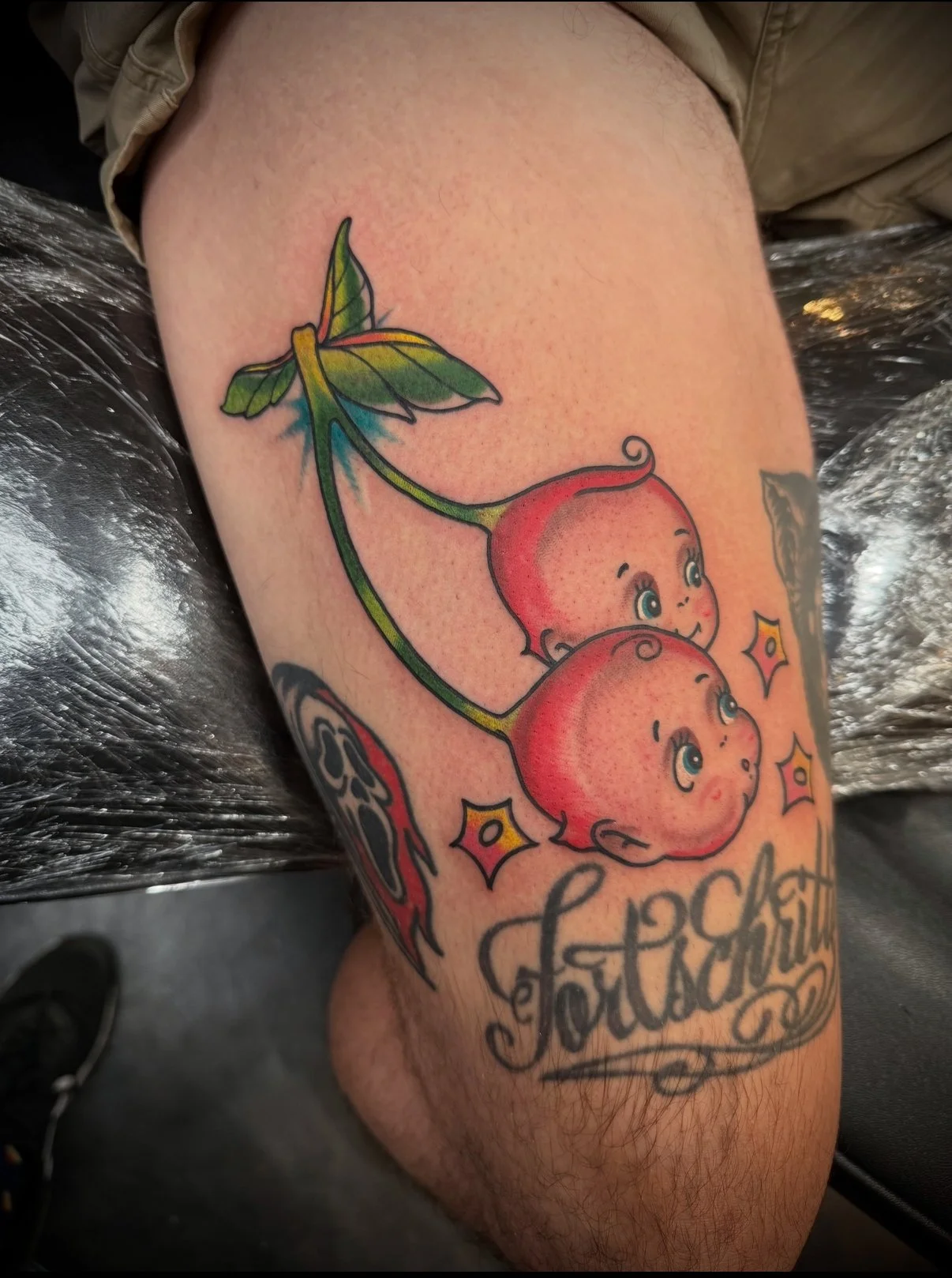 Colorful tattoo of two cherries with smiling kewpie faces, leaves, and stars, with the word 'Fortunate' below.