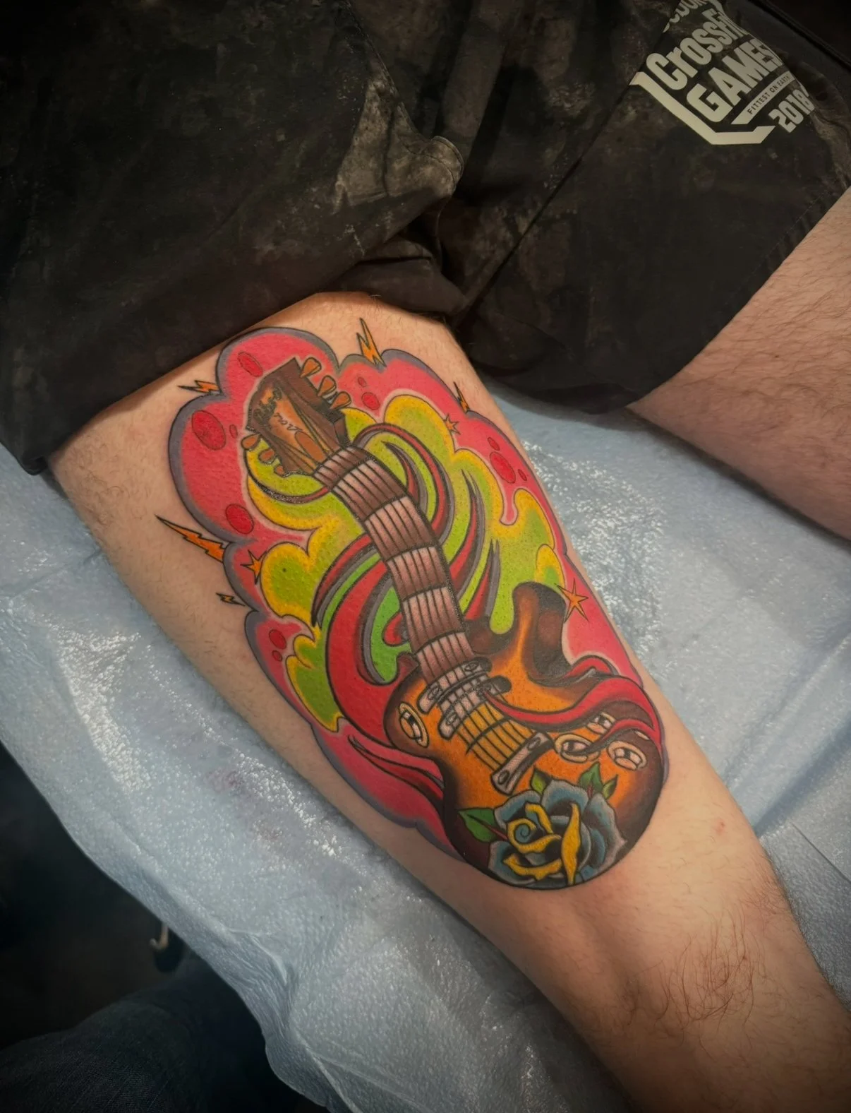 A colorful tattoo of a guitar surrounded by vibrant, swirling colors and lightning bolts on a person's leg.