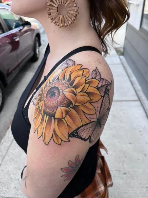 Tattoo of a sunflower with detailed shading and a butterfly on the woman's upper arm.