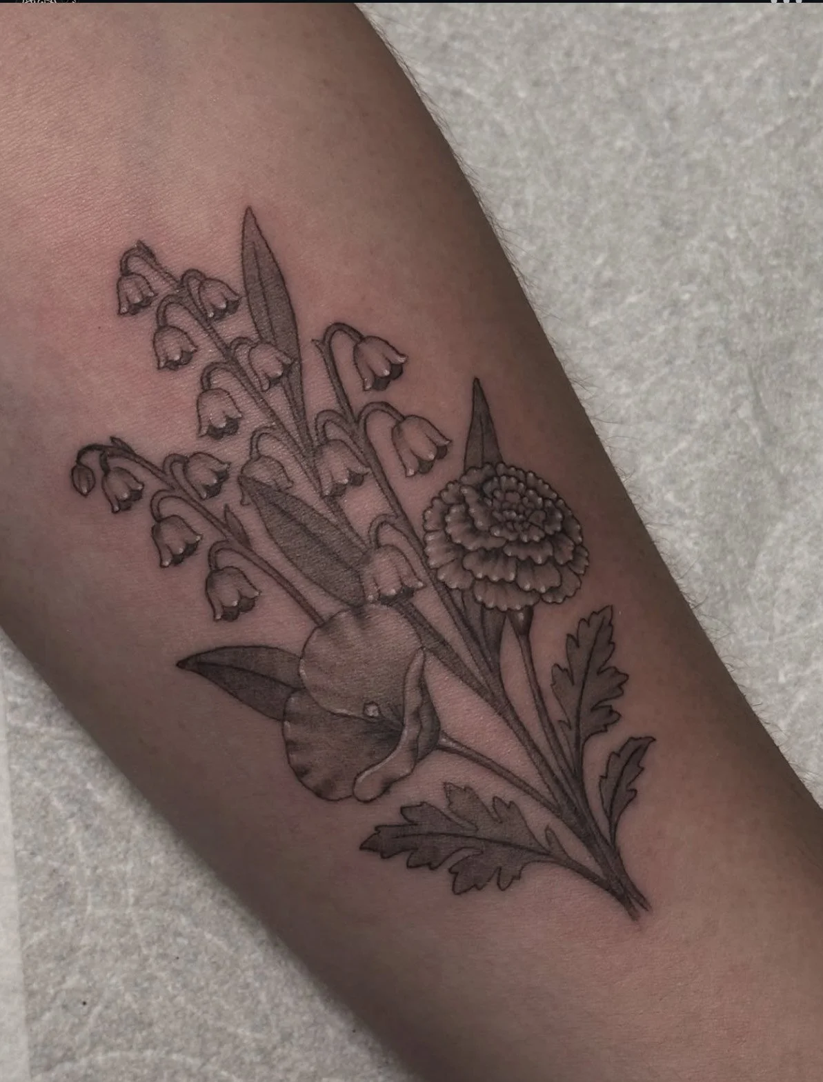 Black fine line outline tattoo of various flowers, including a large marigold, a lily of the valley, and a fern, on a person's arm.