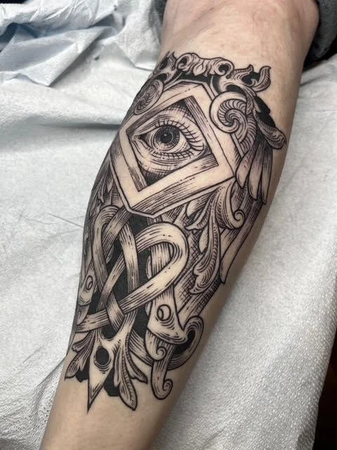 Tattoo of an eye inside a square frame, surrounded by intricate, flowing ornamental designs on a person's forearm.