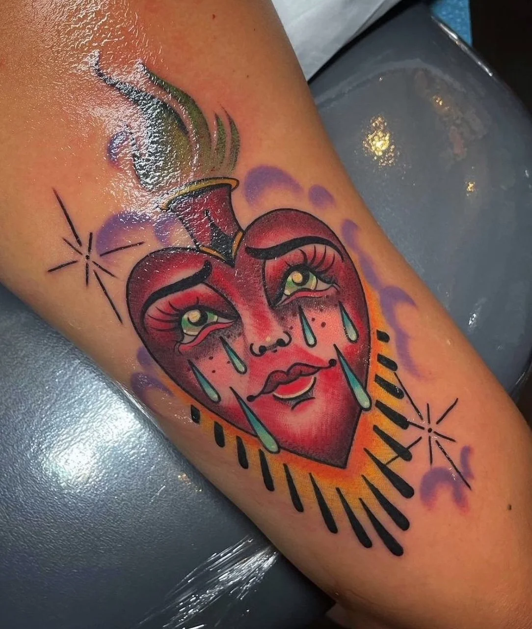 Tattoo of a crying heart with tears, eyes, and a small nose, surrounded by purple clouds and black starburst accents.