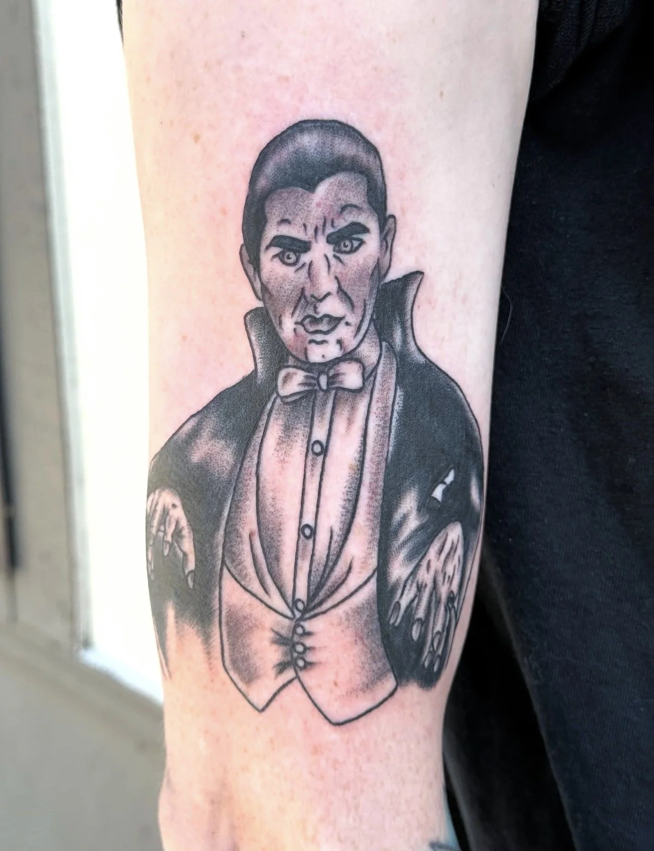 Tattoo of a man with a stern expression, dressed as a vampire, wearing a tuxedo with a bow tie and cape, holding a pair of large, clawed hands