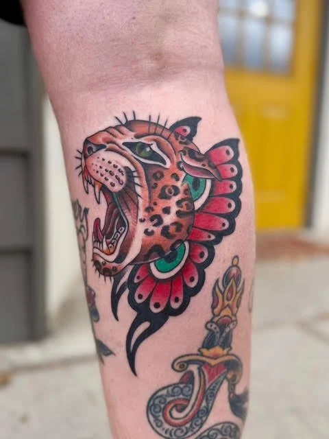 Colorful traditional tattoo of a leopard's head with black spots and green eyes, incorporated into a floral design on a person's leg.