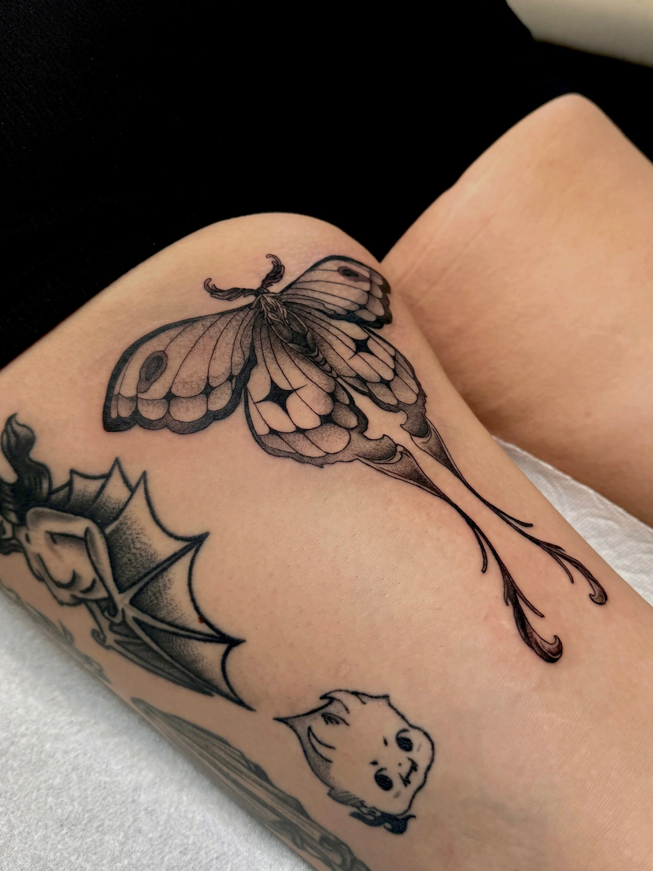Tattoo of a butterfly, with a spider web and a small animal face, on a person's thigh.