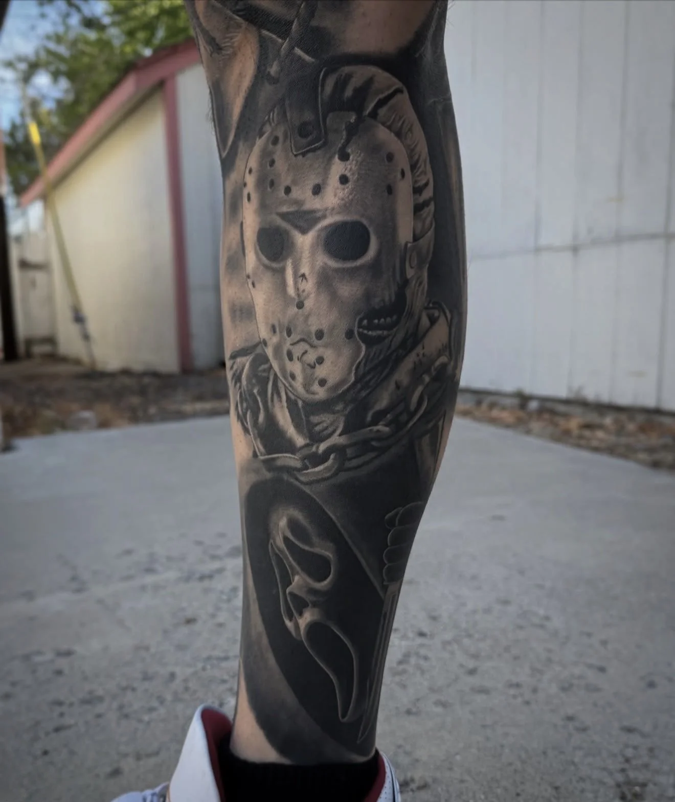 Black and gray realism tattoo of a jason mask and a scream mask, featuring chains, on a person's forearm.
