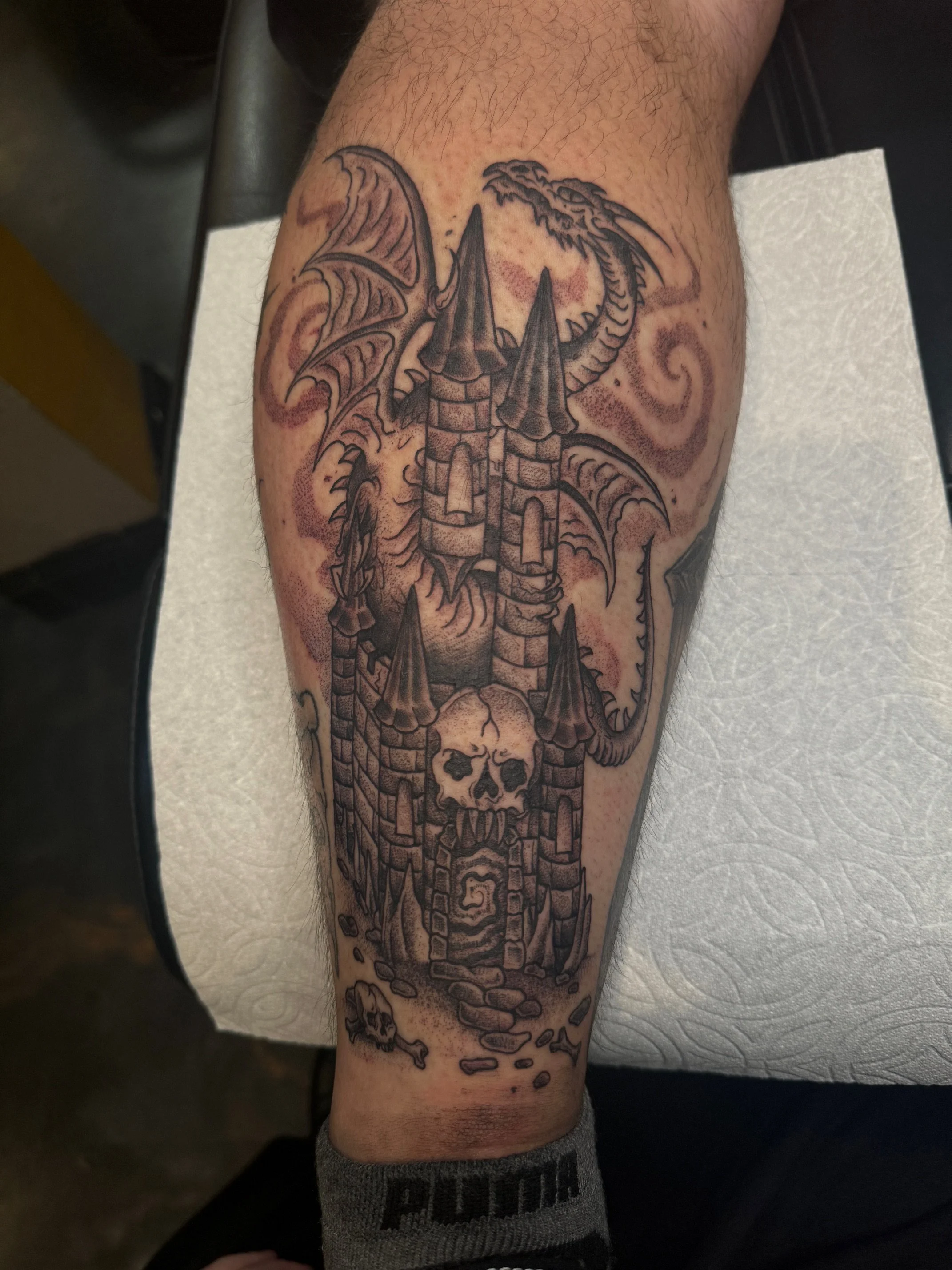 Tattoo of a castle with skulls, dragons, and clouds on a person's leg.