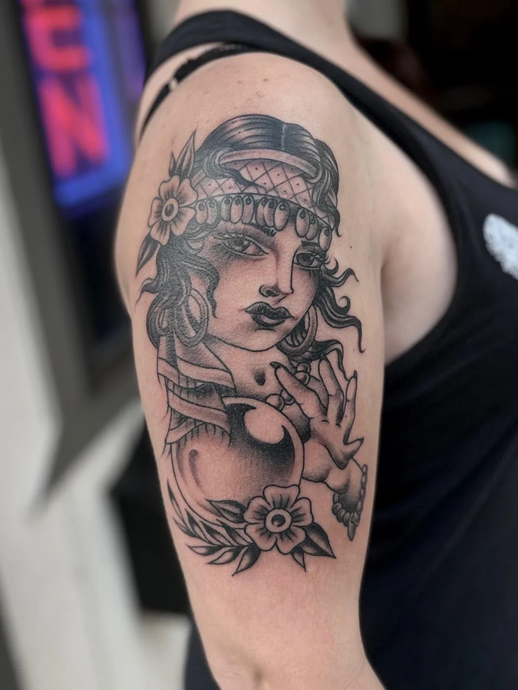 Tattoo of a woman with long hair, flowers, and jewelry on upper arm, in black and gray ink.