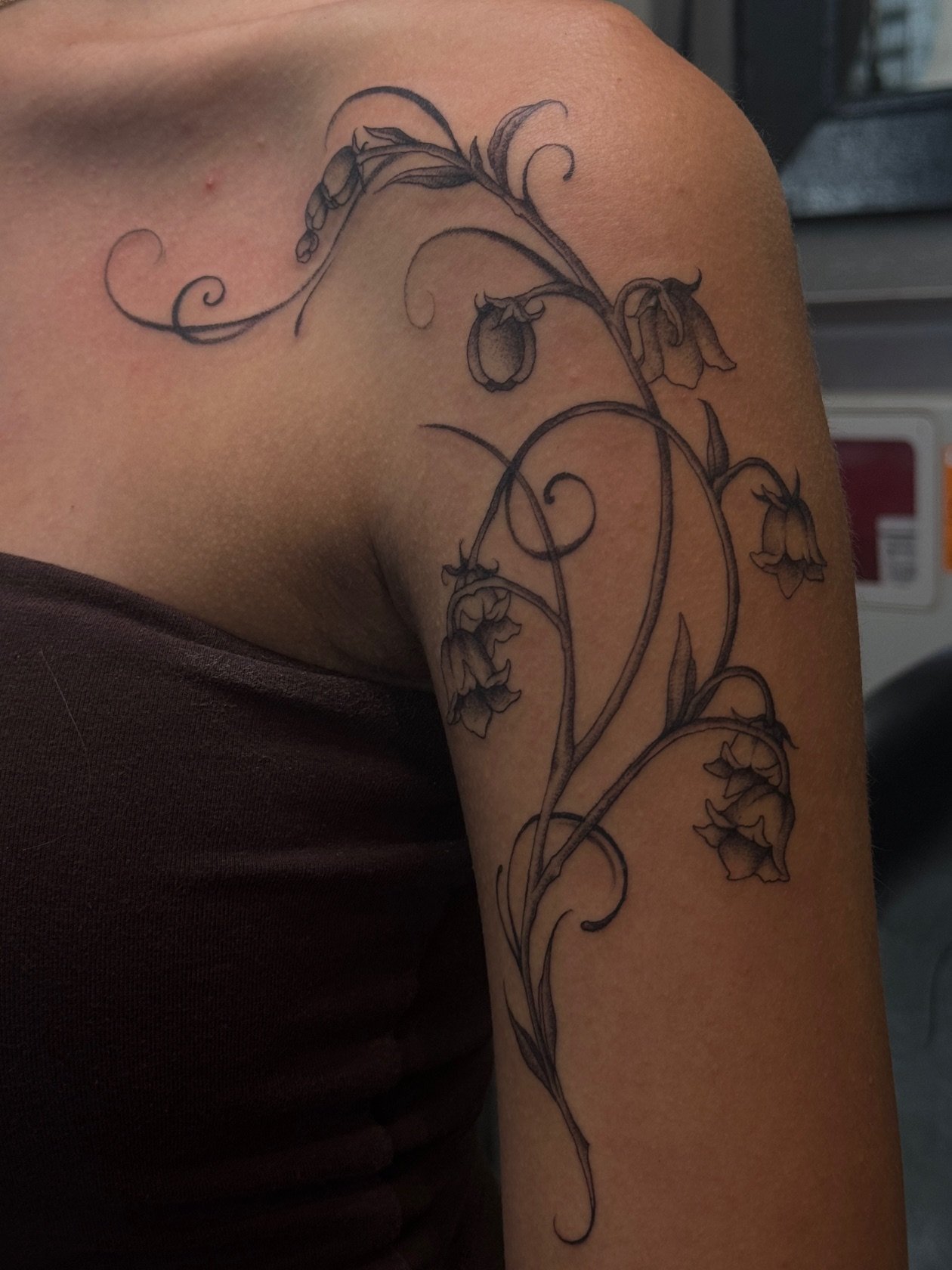Tattoo of a floral design with bell-shaped flowers and curling vines on a person's upper arm and shoulder.